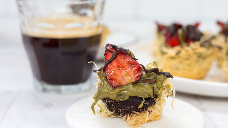 Half of a Dubai chocolate strawberry cup on small plate with coffee and more treats in background