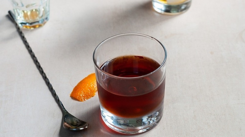 Manhattan cocktail in glass next to cocktail stirrer and slice of orange peel