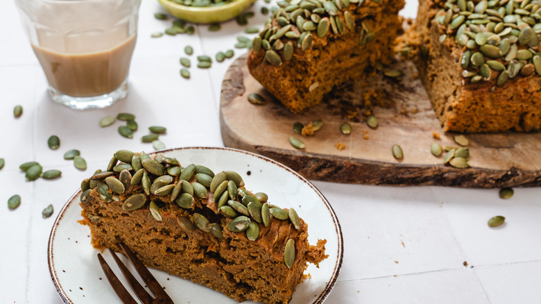 A slice of pumpkin bread on small plate next to sliced pumpkin bread on a cutting board next to cup of coffee