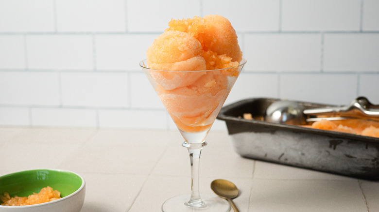 A glass of cantaloupe sorbet sits beside a spoon, a pinch bowl with diced cantaloupe and a container of ice cream in the back
