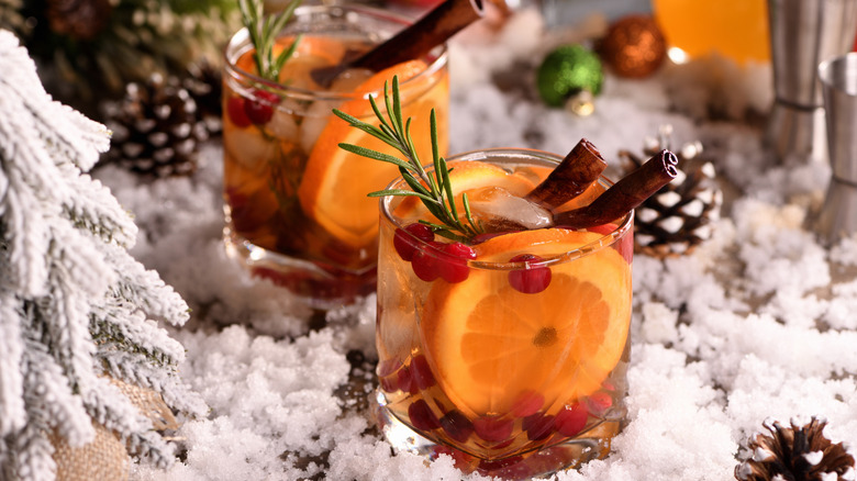 Glasses on whiskey with orange, cranberries, cinnamon sticks, rosemary sprig on white snowy surface