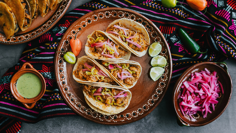Tacos with pickled onions on a plate beside lime wedges and chiles atop a colorful cloh