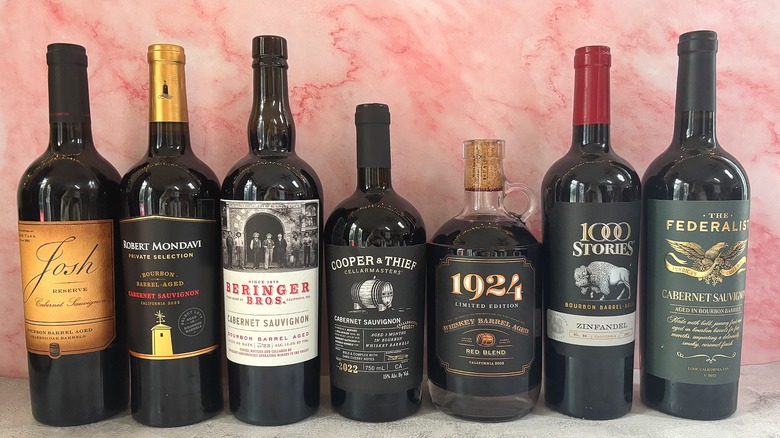 Seven bourbon barrel aged wine bottles lined up on pink background