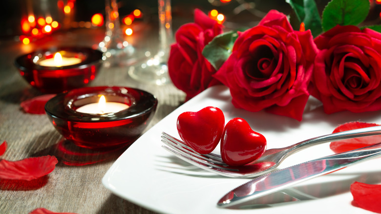 Valentine's theme dinner table close-up with 2 red hearts on a fork balanced on a white plate with red roses and tea lights on the table