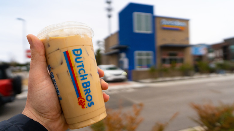 Hand holding a Dutch Bros iced coffee with the drive-thru blurred in the background