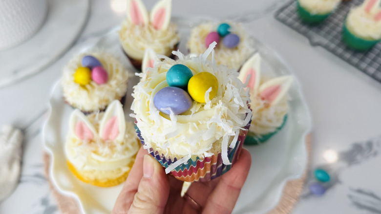 hand holding a white-frosted cupcake topped with coconut and three candy eggs