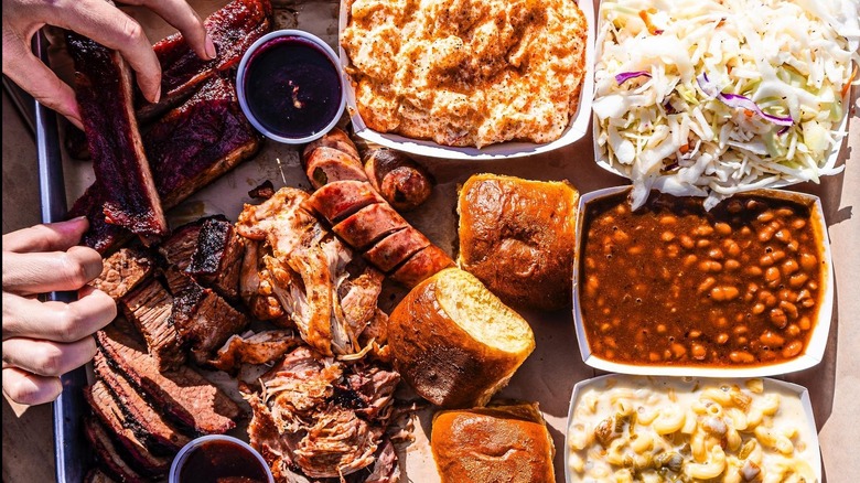 Hand grabbing ribs from a barbecue platter consisting of ribs, sausage, pulled meat, mac and cheese, cole slaw, baked beans, rolls, and more