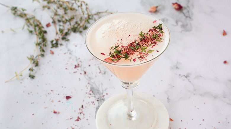Blush pink cocktail in martini glass, garnished with crushed rose petals and thyme leaves