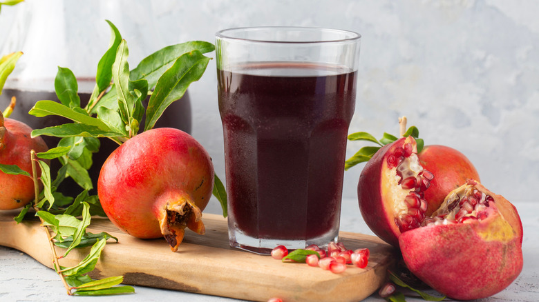 A large glass of pomegranate juice next to whole and torn fruits on wooden cutting board.