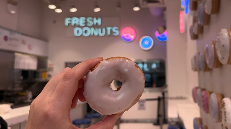 person holding up a gluten-free donut with a sign that says Fresh Donuts behind it
