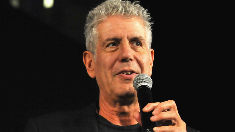 Anthony Bourdain speaking into a microphone dressed in a black blazer and t-shirt