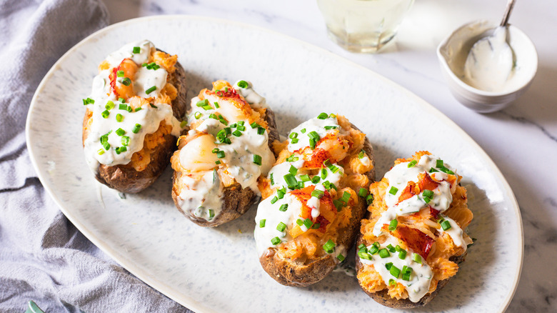 Four lobster-stuffed baked potatoes on a platter with sauce, chives, and glass of wine.