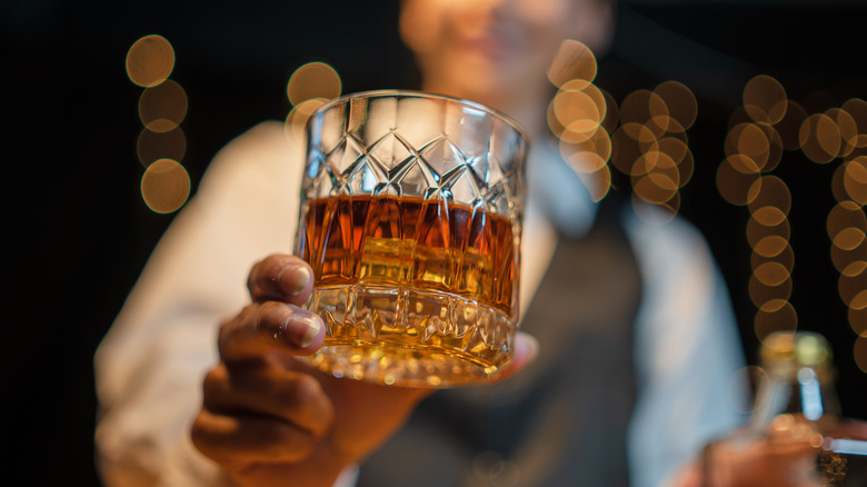 A tumbler of whisky being held out toward the camera by a blurred bartender