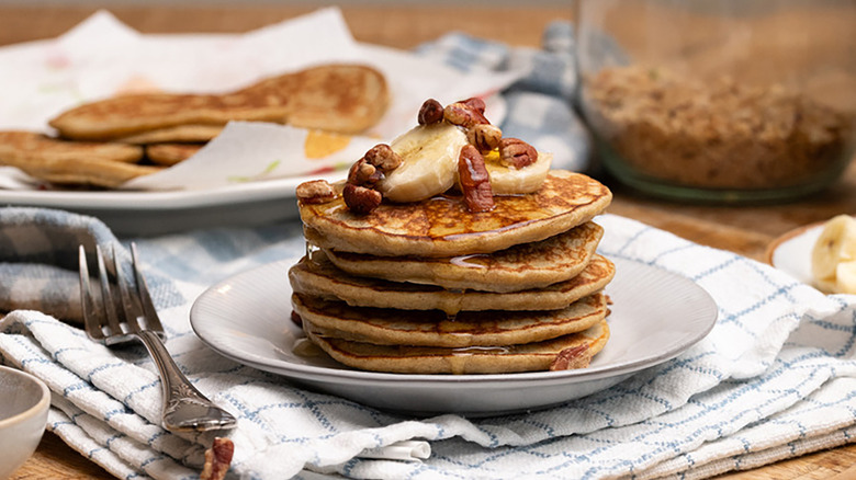 Stack of banana pancakes on plate, topped with chopped pecans and sliced banana