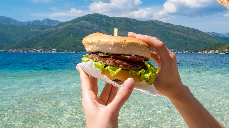 Hands holding a burger in front of a seaside view.