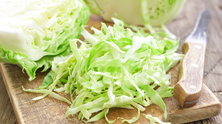 A head of cabbage on a wooden cutting board, next to shredded cabbage and a knife