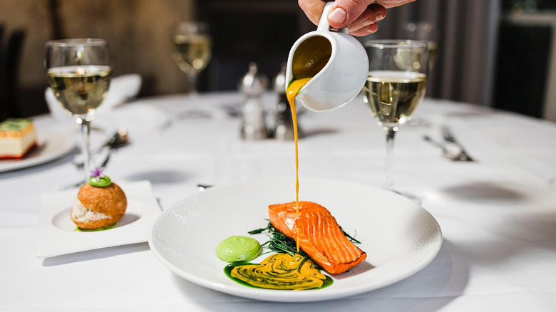 Hand pouring sauce over a salmon dish on a white tabletop with a formal setting