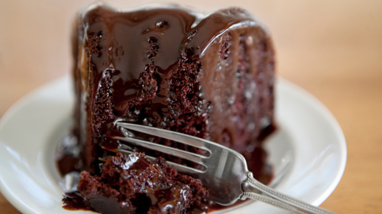A piece of decadent chocolate cake is covered in chocolate sauce on a plate.