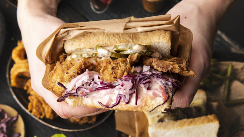 A person holds a condiment-loaded fried chicken sandwich wrapped in brown paper