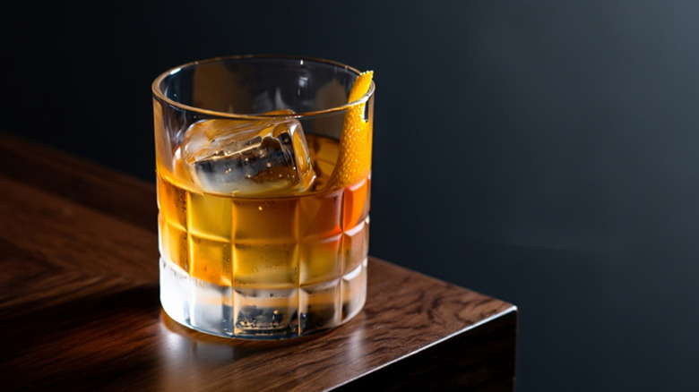 An Old Fashioned cocktail with ice and orange zest sitting at the corner of a dark wooden table against a black background