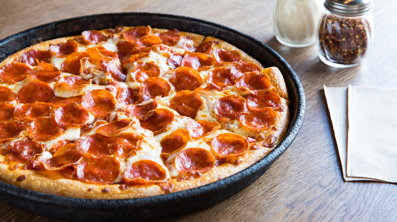 A Pizza Hut pepperoni pizza still in the pan sitting on the table next to shakers of red pepper and Parmesan