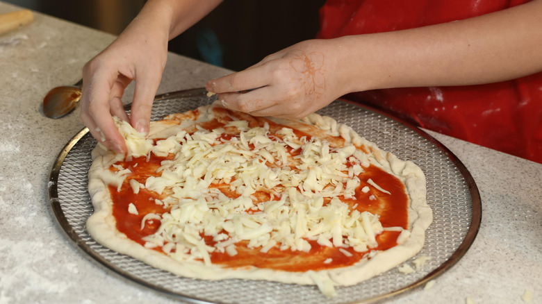 A person in red sprinkles shredded mozzarella cheese on pizza that hasn't been baked