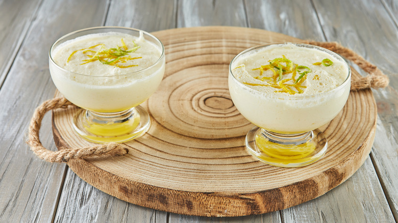 Two glass bowls of lemon mousse topped with citrus zest, resting on a wood platter