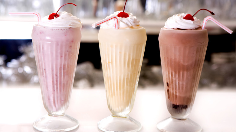 A restaurant countertop with three milkshakes (strawberry, vanilla, and chocolate) in frosted glasses, topped with whipped cream and a cherry