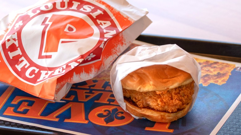 Fried chicken sandwich from Popeyes wrapped in white paper on tray next to red and orange logo paper bag.