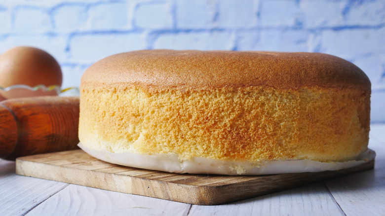 Whole round shaped baked yellow cake with wax paper underneath sitting on top of wooden board.