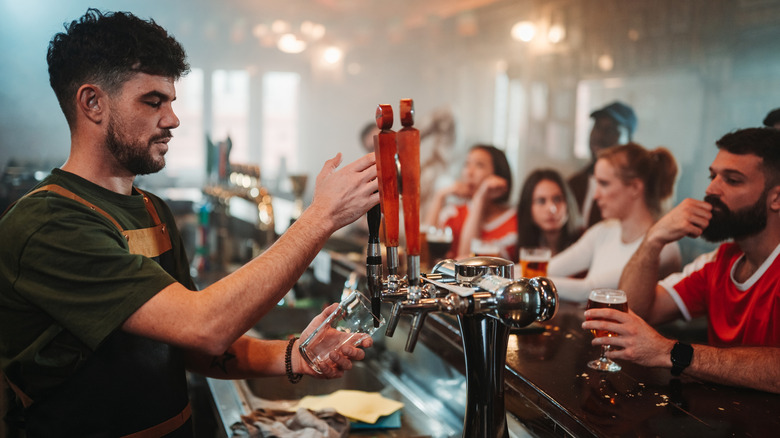 A bartender pours a pint of beer while a number of customers sit at the bar in front of him