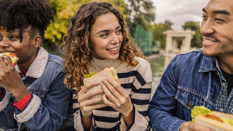 Three people sitting together outside smiling and eating sandwiches