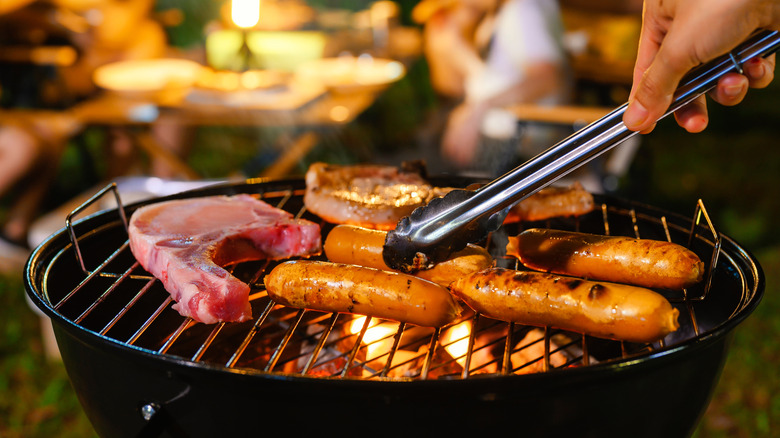 Person using tongs to move sausages on flaming barbecue grill next to bone in steak and pork chops