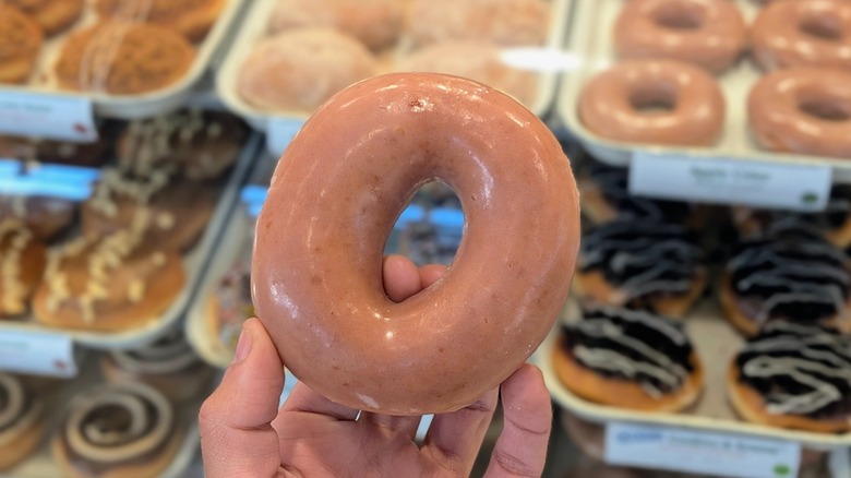 A hand holding an Apple Cider Original Glazed donut in front of the pastry case at Krispy Kreme
