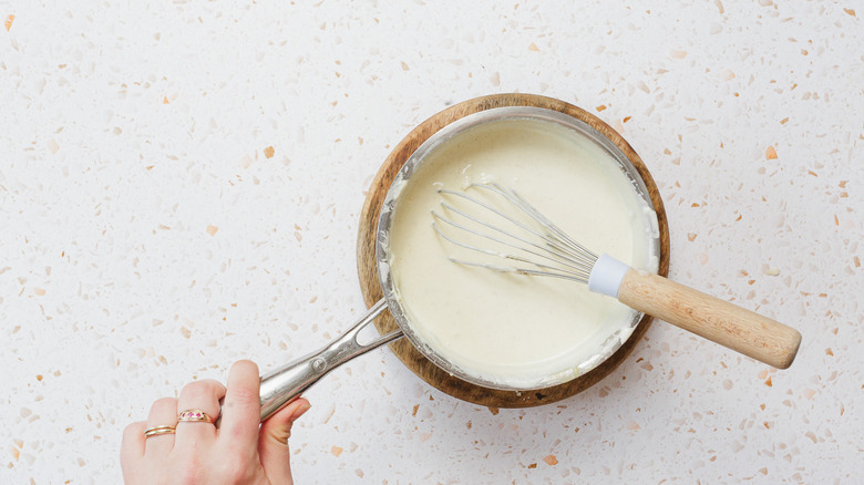 Hand holding saucepan with creamy sauce and whisk