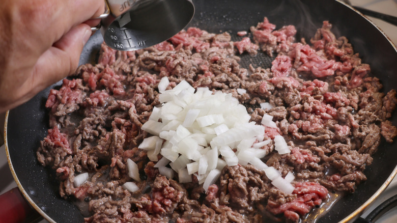 Hand pouring measuring cup of diced onion into skillet of ground beef