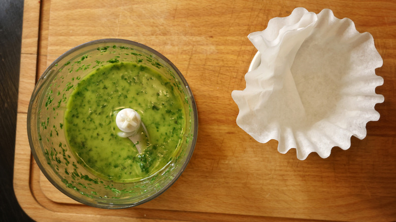 food processor with pureed basil oil and coffee filter next to it on cutting board