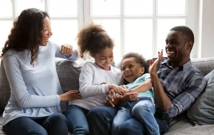 Happy black family laughing playing with children at home, cheerful african american parents tickling 2 mixed race kids having fun on couch, mom dad with little son daughter enjoying funny activity
