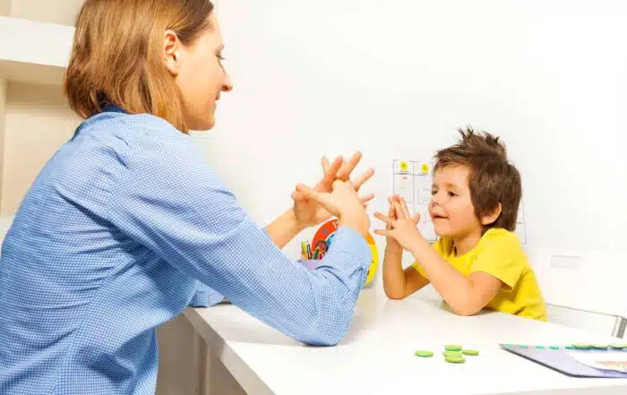 Boy exercises by putting hands and fingers together with therapist showing it improving motor skills sitting opposite at the table indoors