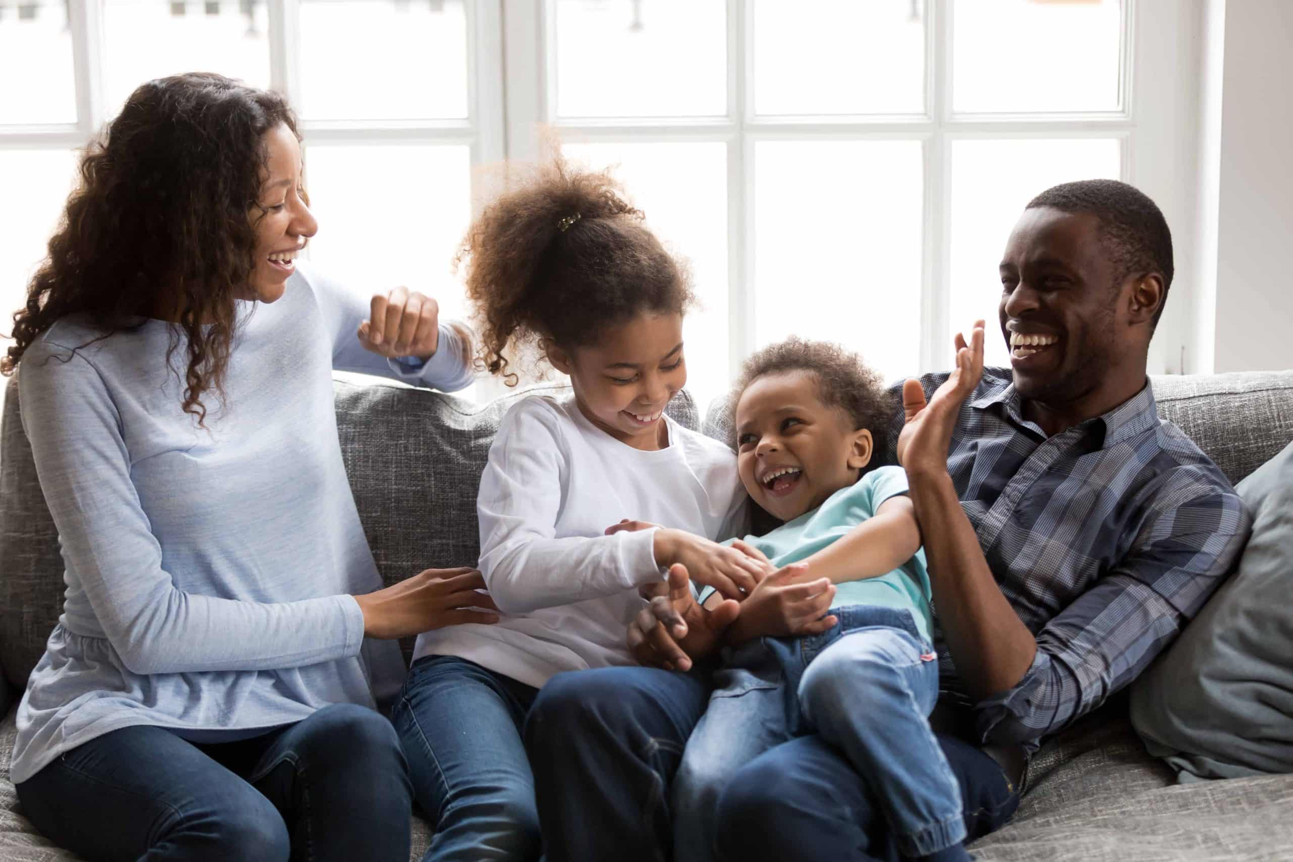 Happy black family laughing playing with children at home, cheerful african american parents tickling 2 mixed race kids having fun on couch, mom dad with little son daughter enjoying funny activity