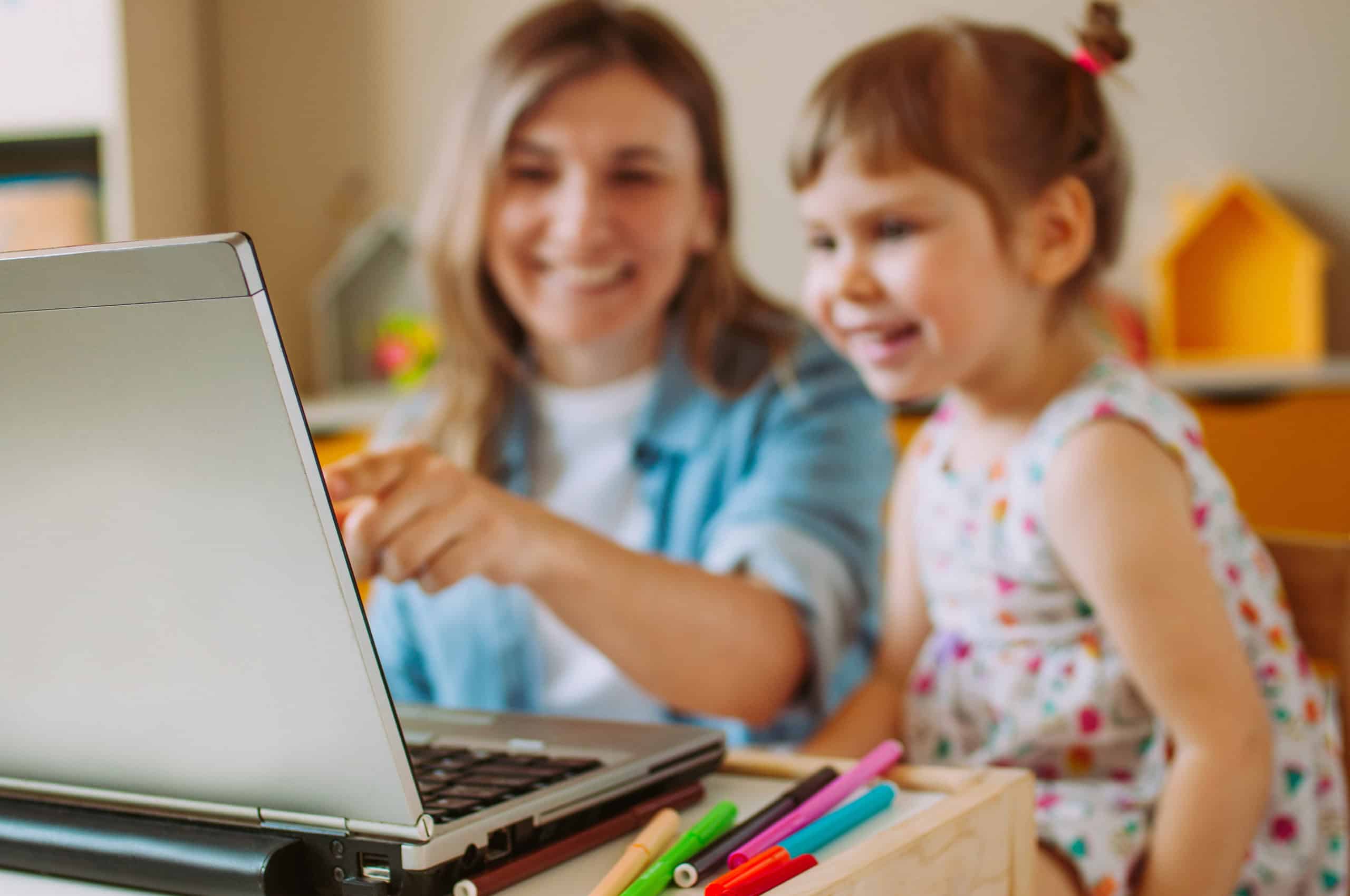 Smiling mother and toddler daughter looking at the notebook indoor. Distance online learning concept. Activities for kids during quarantine. Focus on the notebook.