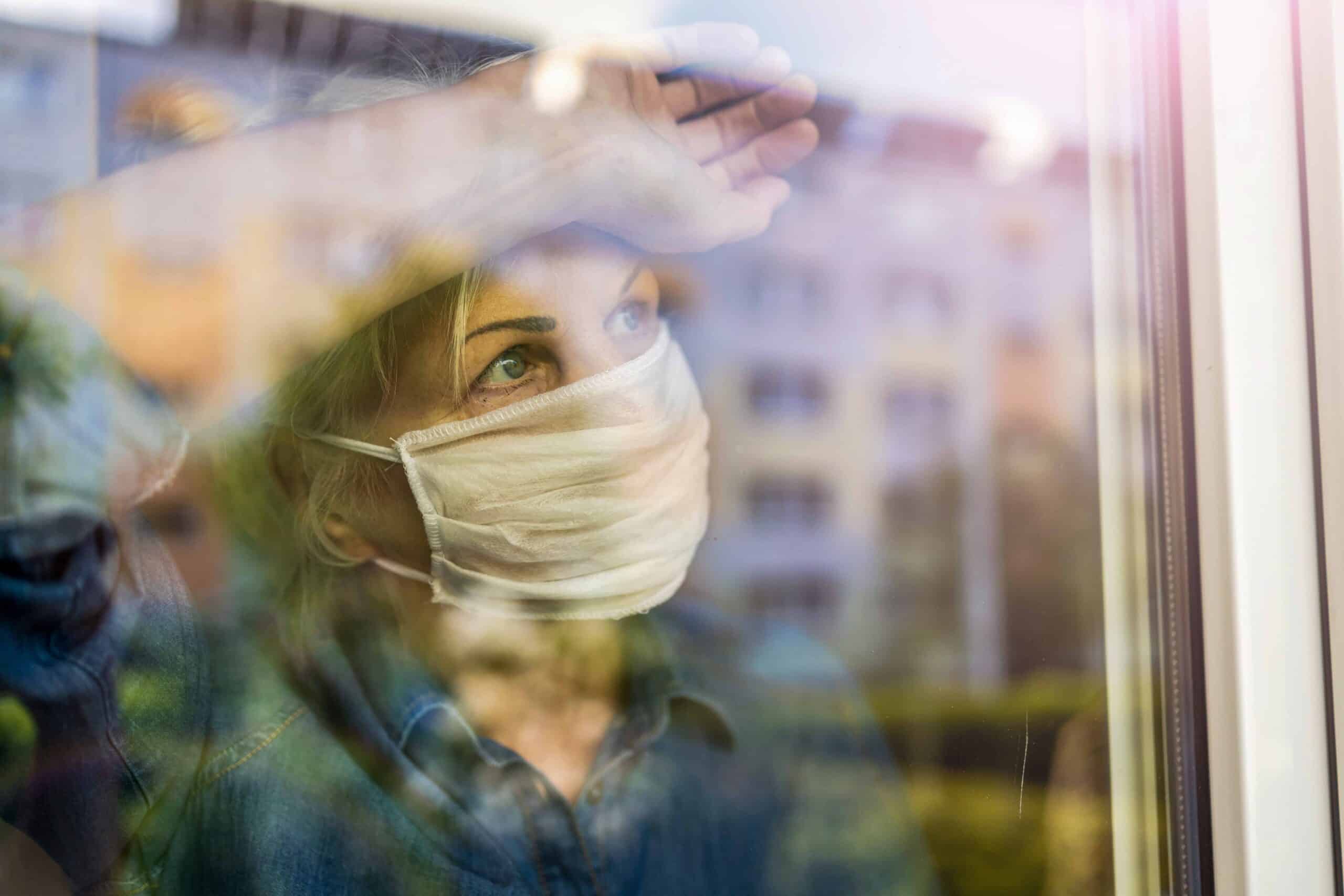 Close-up of a woman wearing a white mask, looking outside through a window