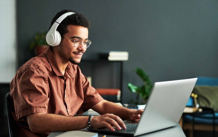 Young smiling man in headphones typing on laptop keyboard while sitting by workplace and taking part in online webinar or lesson