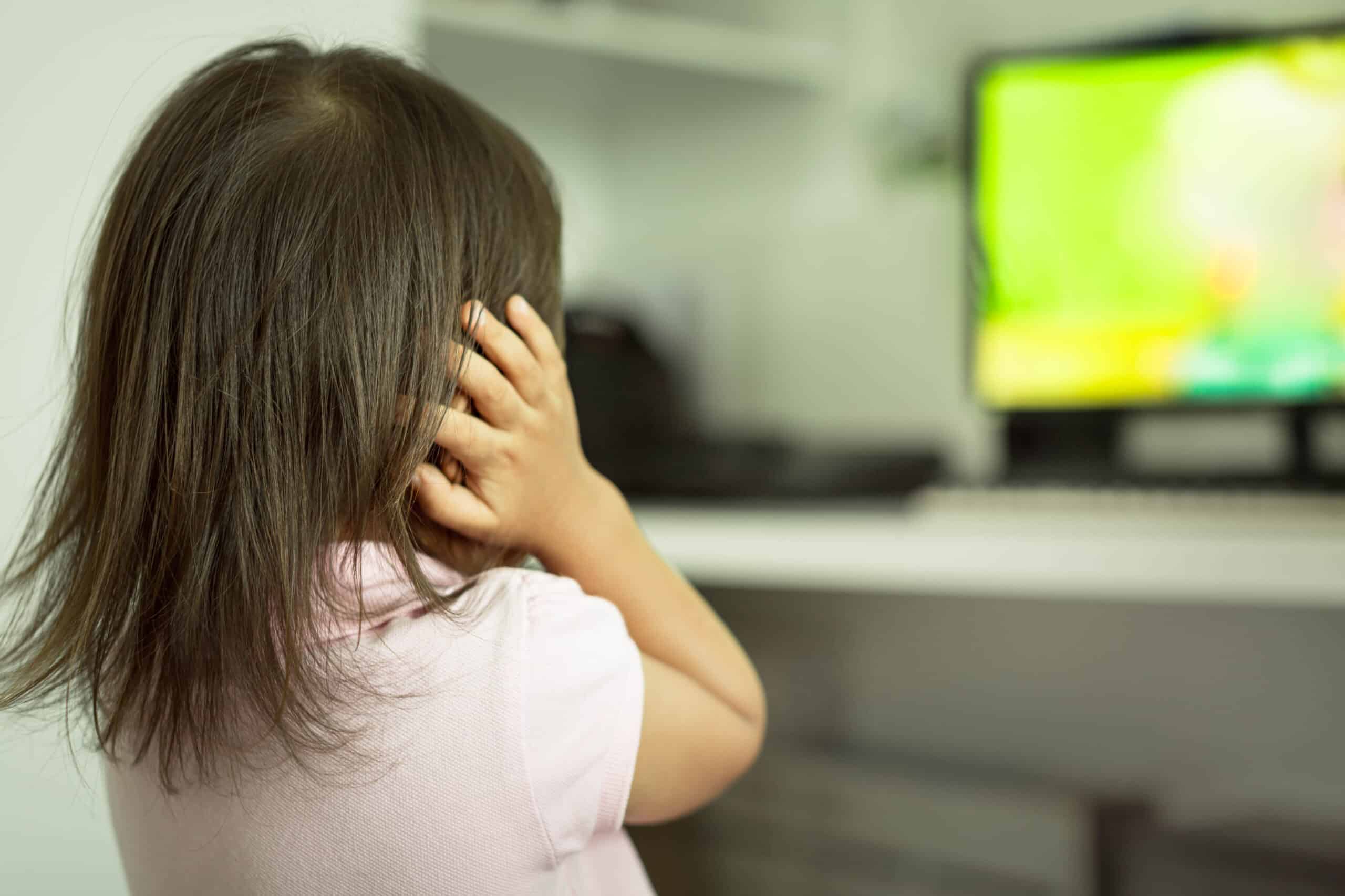 Child covering her ears while looking at a TV screen in the background