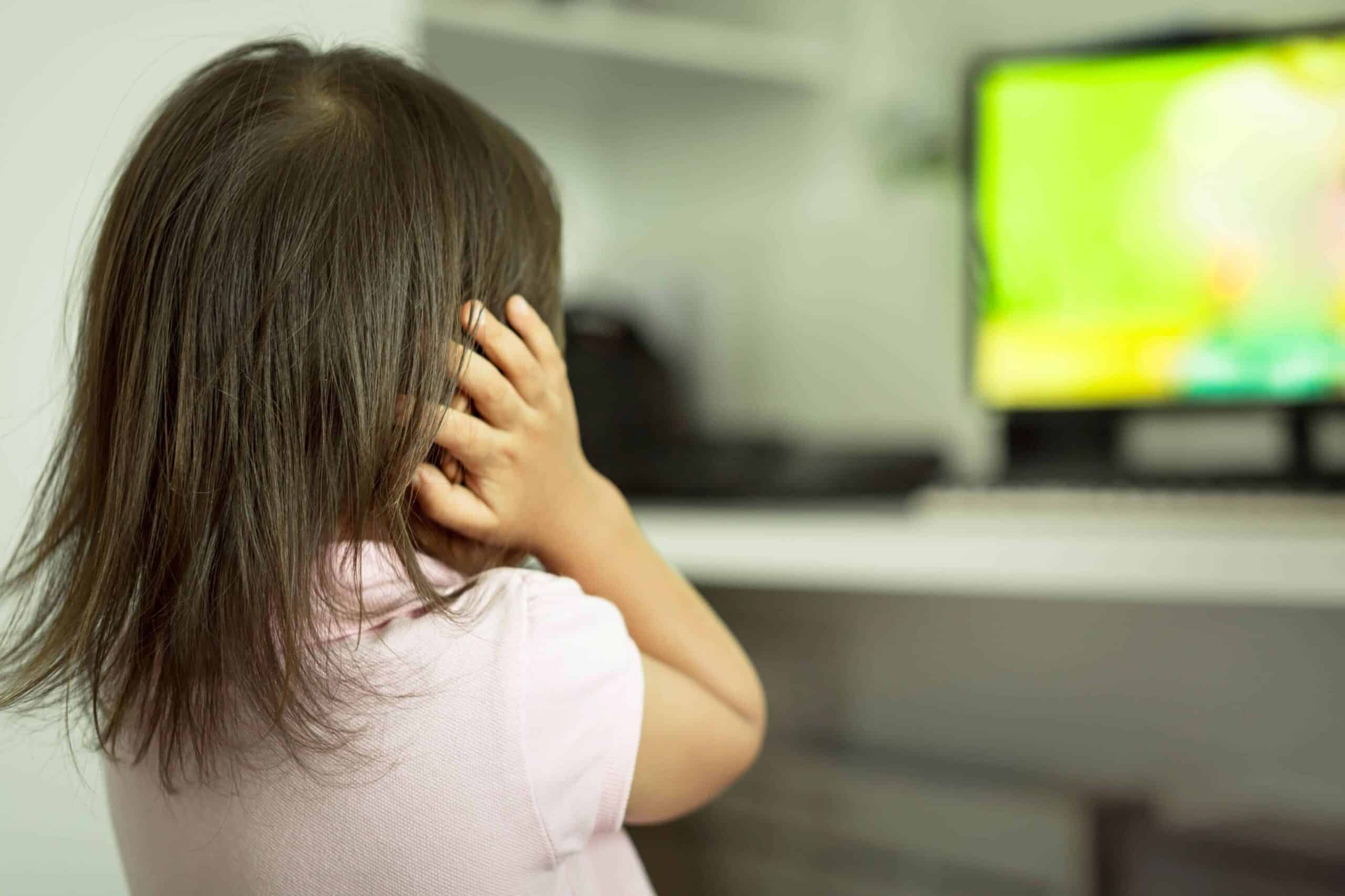 Child covering her ears while looking at a TV screen in the background