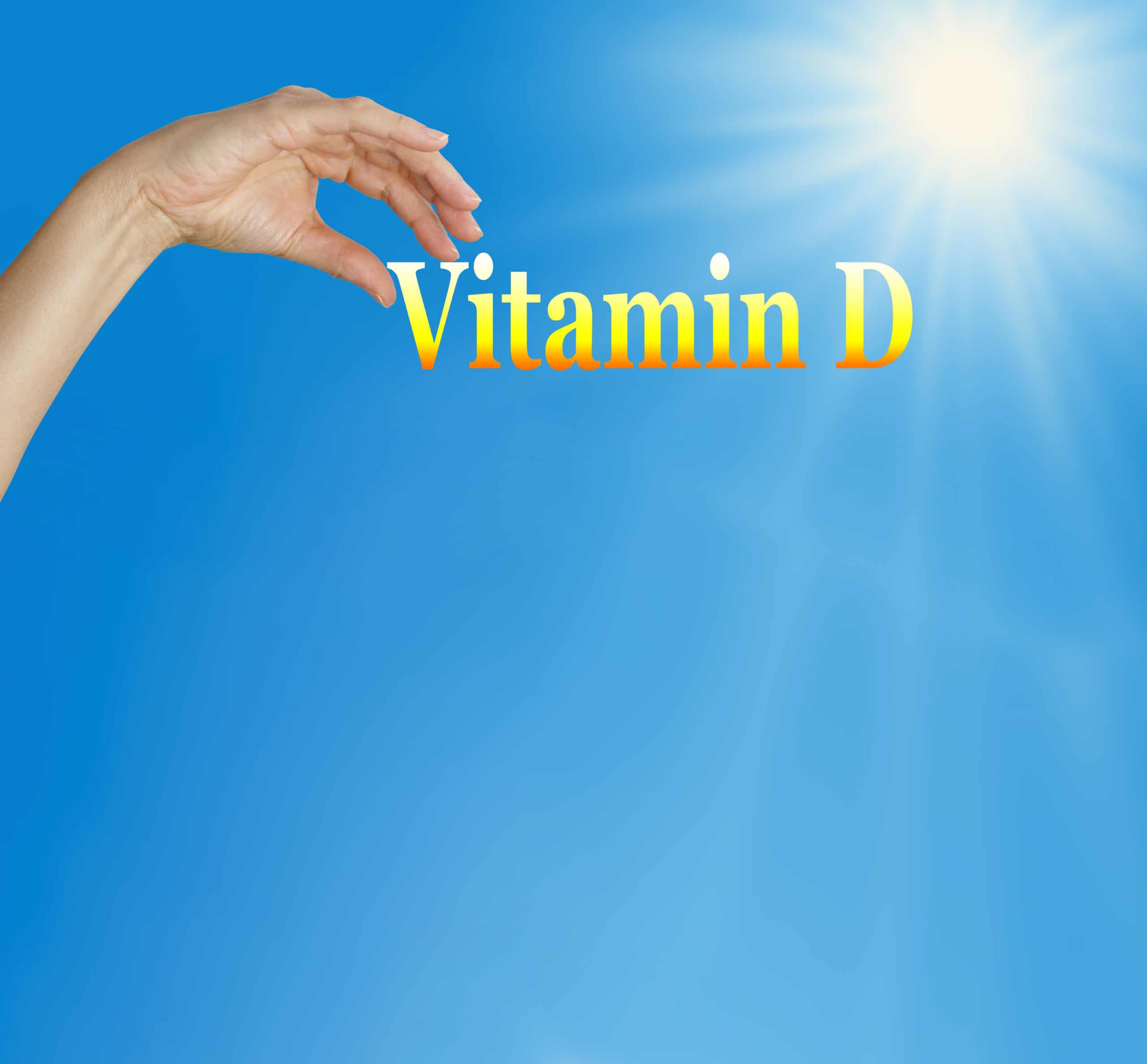 Hand holding up the words "vitamin D" on blue sky background with sunshine coming from upper right.